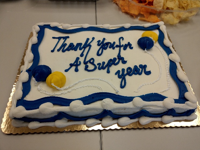 A cake with white icing and blue trip with yellow ballons. The cake says 'Thank you for a super year.'